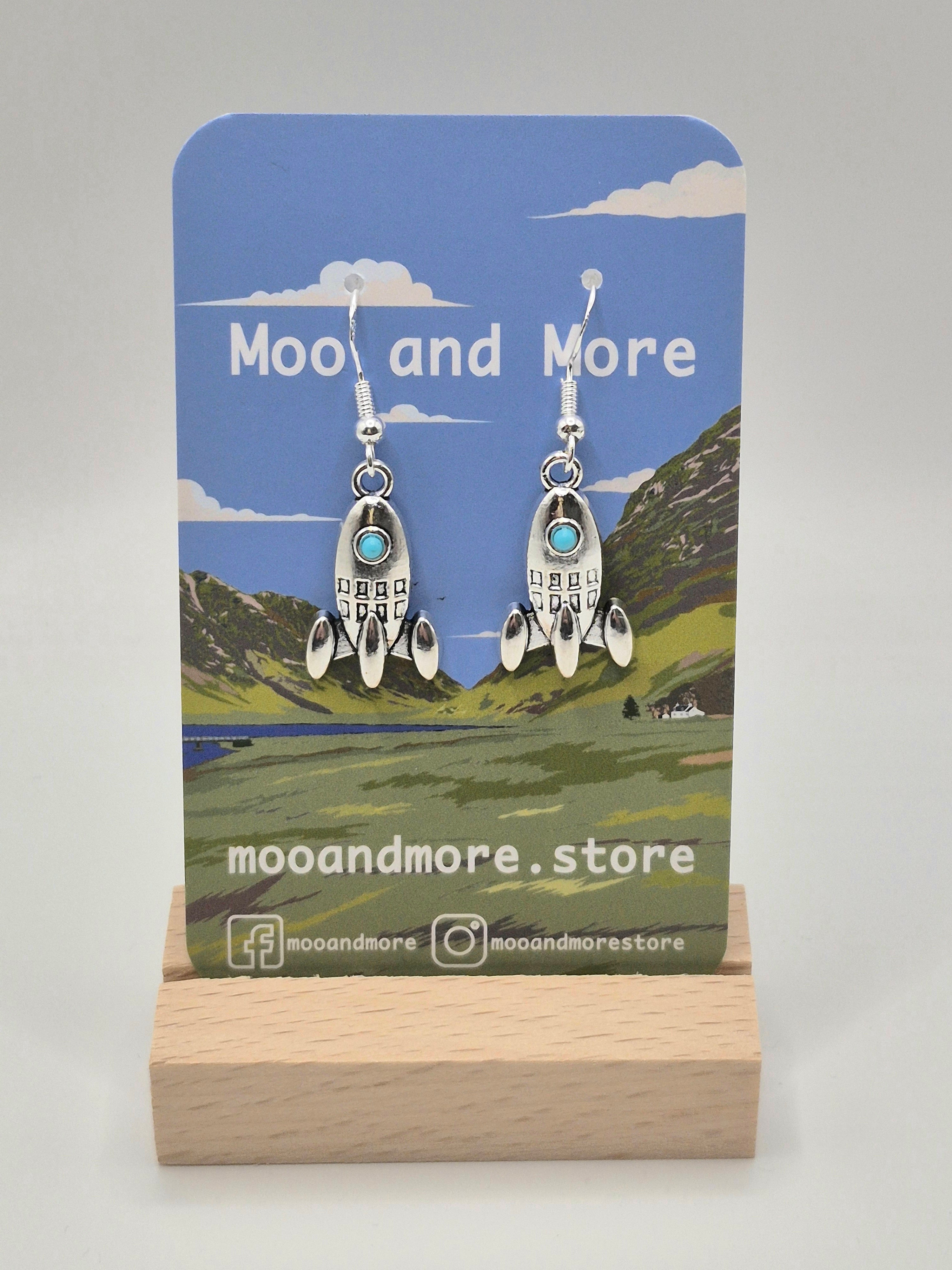 A pair of sterling silver rocket-shaped dangle earrings with small turquoise blue windows, displayed on a clear plastic hanger against a light gray background.