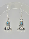 A pair of sterling silver rocket-shaped dangle earrings with small turquoise blue windows, displayed on a clear plastic hanger against a light gray background.