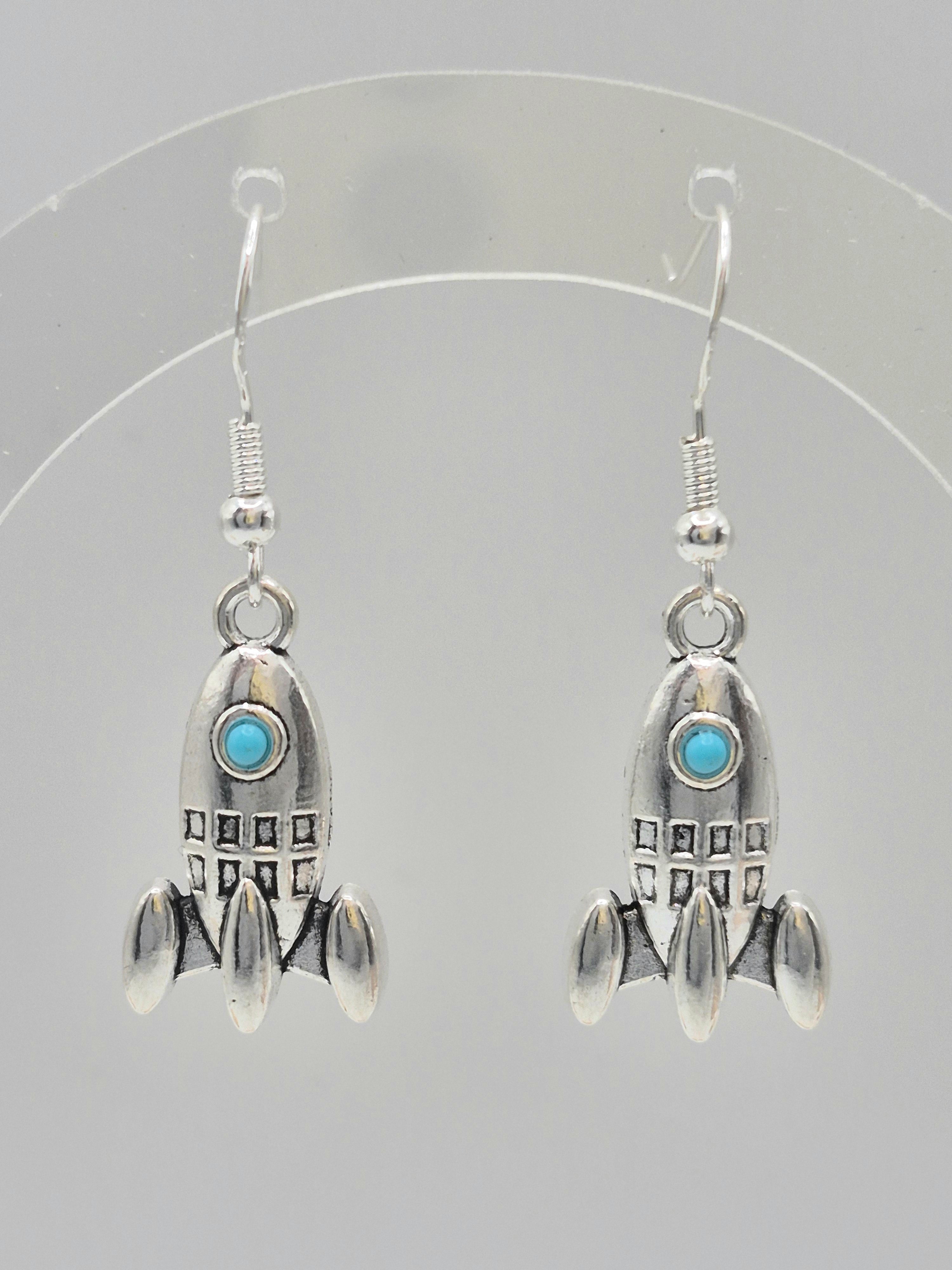 A pair of sterling silver rocket-shaped dangle earrings with small turquoise blue windows, displayed on a clear plastic hanger against a light gray background.
