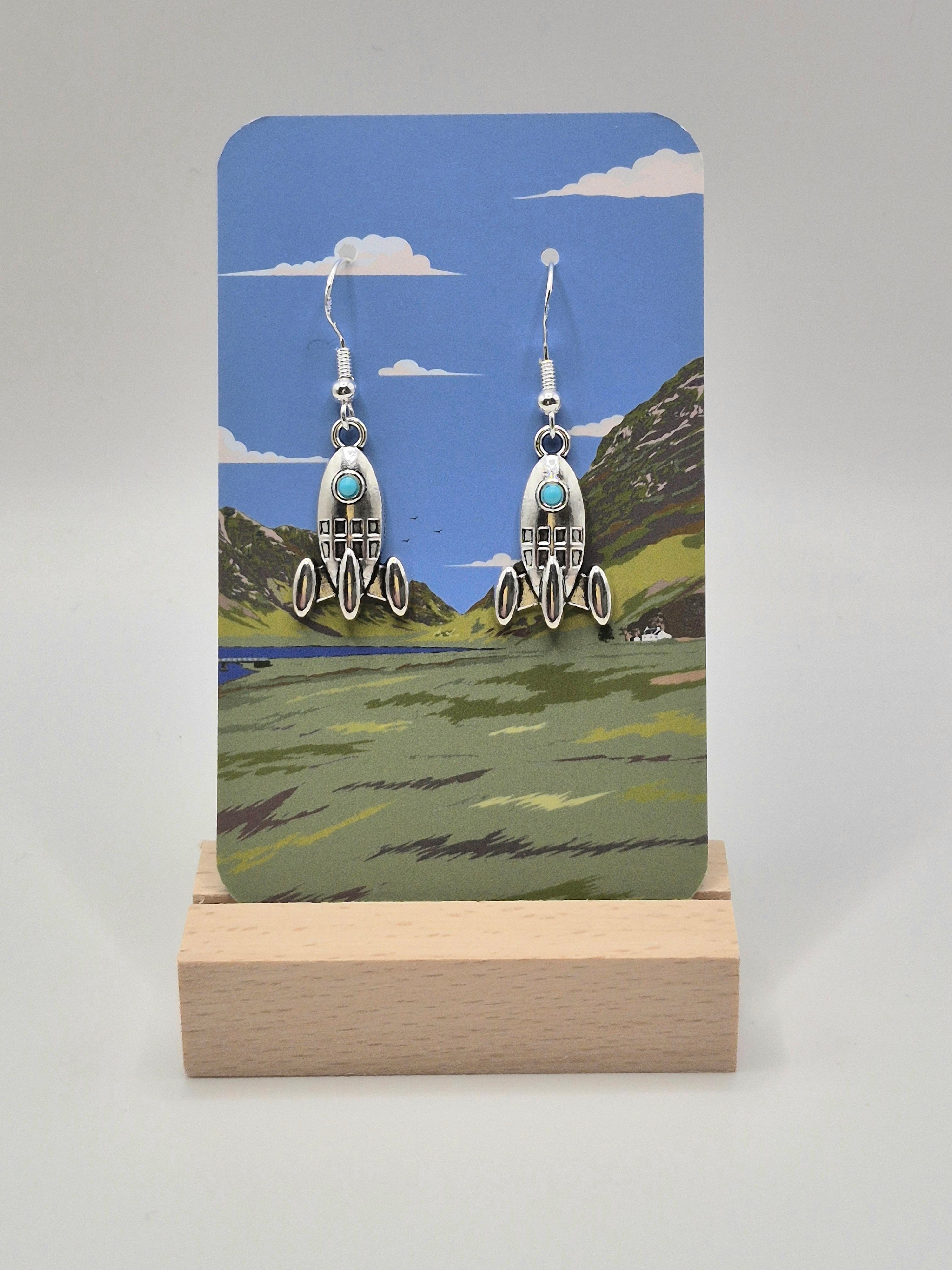 A pair of sterling silver rocket-shaped dangle earrings with small turquoise blue windows, displayed on a clear plastic hanger against a light gray background.