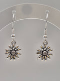 A pair of silver-toned daisy drop earrings with textured centers and hook backings, displayed on a clear circular stand against a neutral gray background.