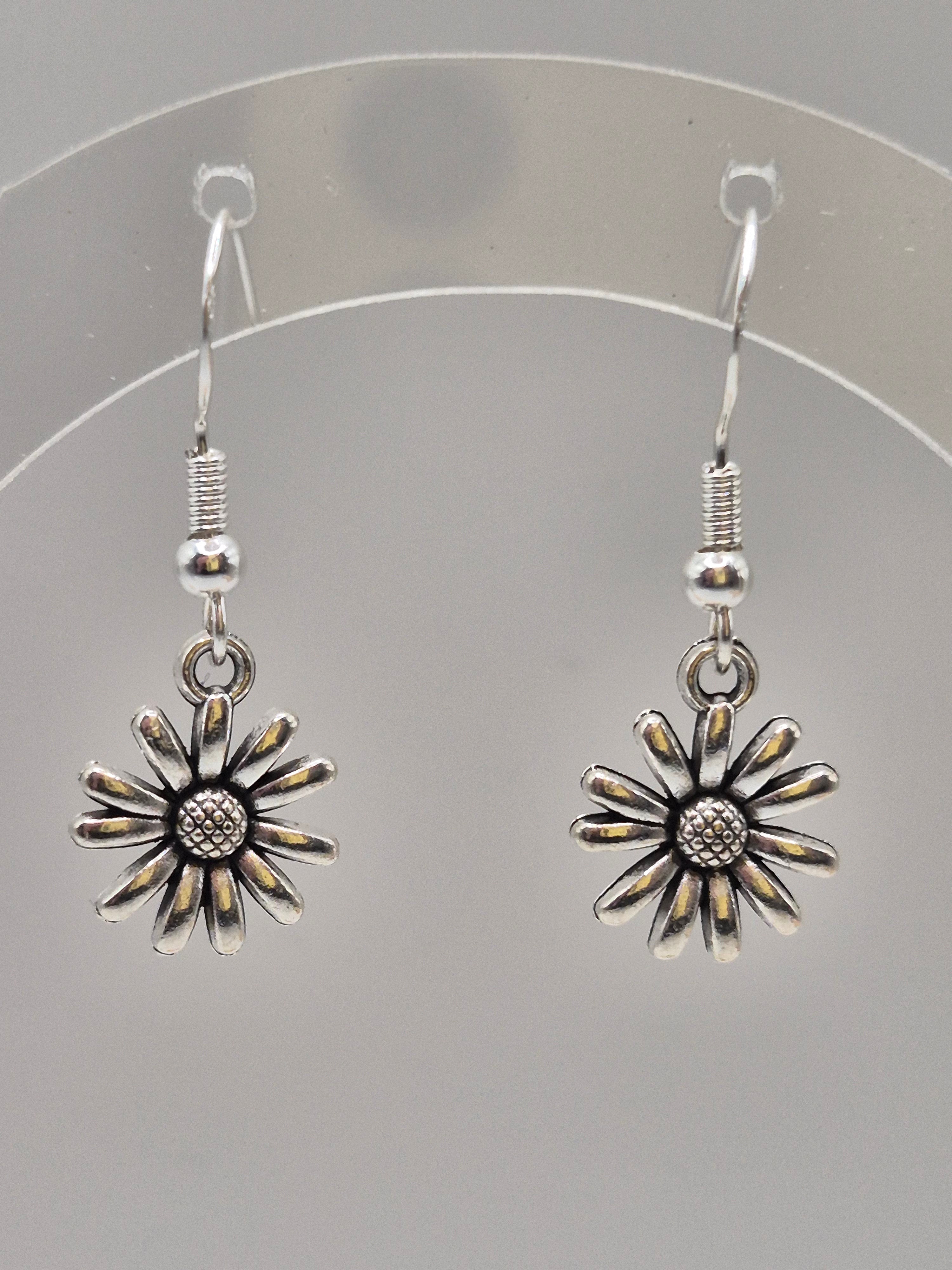 A pair of silver-toned daisy drop earrings with textured centers and hook backings, displayed on a clear circular stand against a neutral gray background.