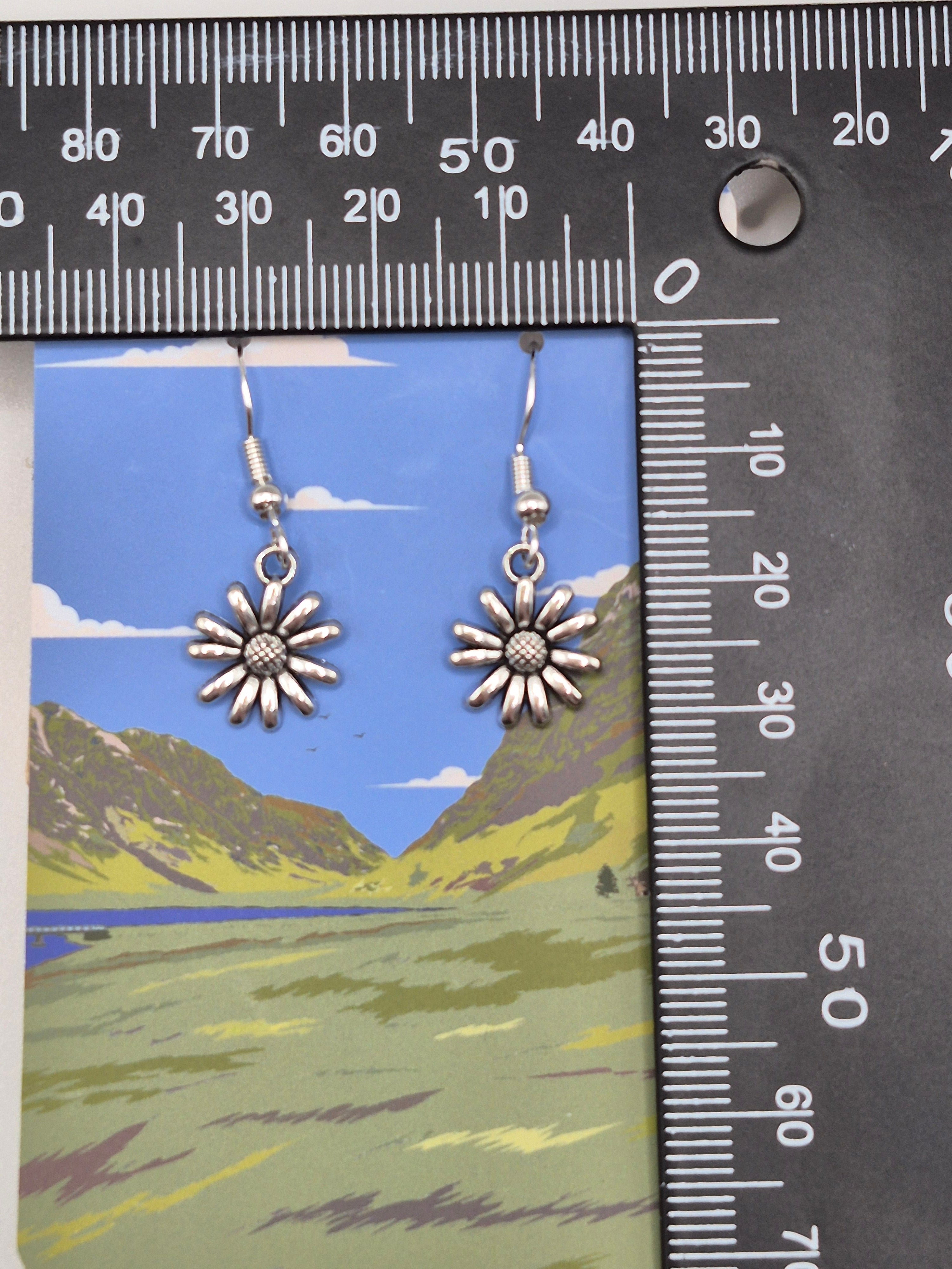 A pair of silver daisy flower drop earrings displayed on a colorful card, with a black metal ruler showing centimeter measurements along the top and left side for scale.