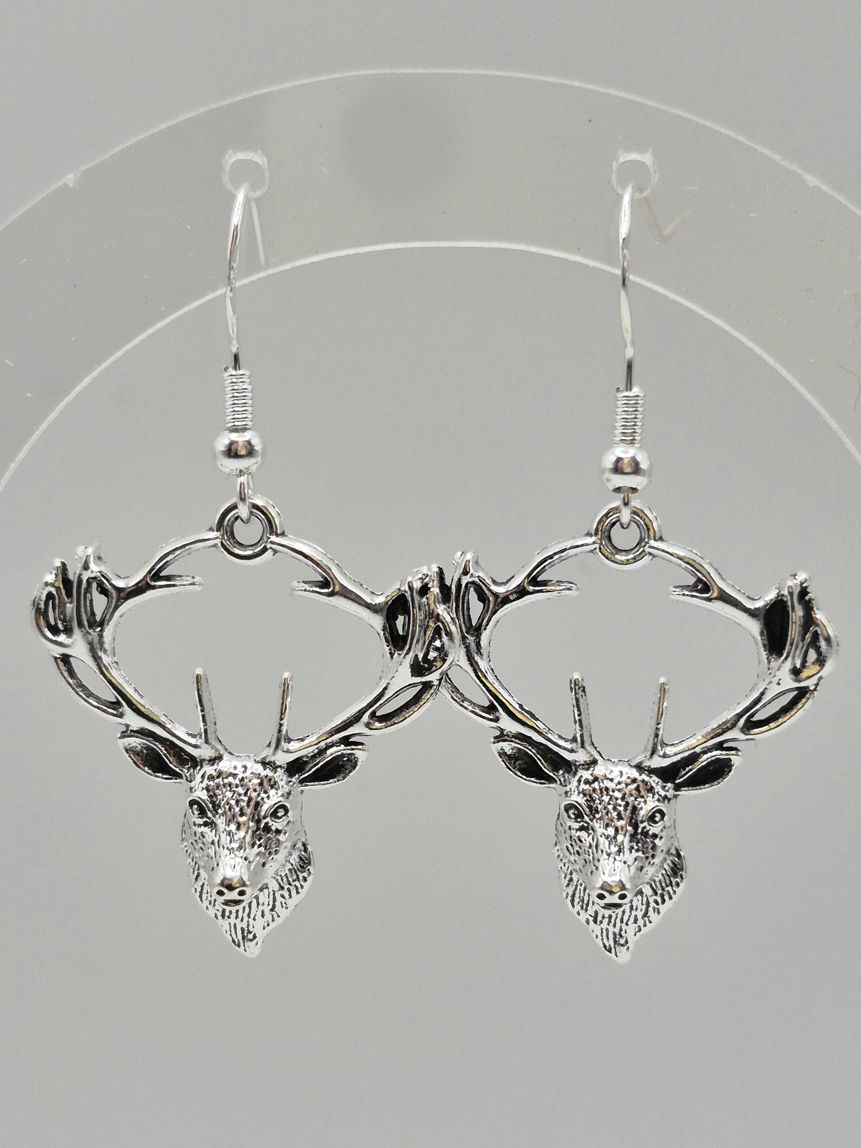 A pair of silver-toned dangle earrings featuring detailed stag head and antler designs, displayed against a neutral gray background.